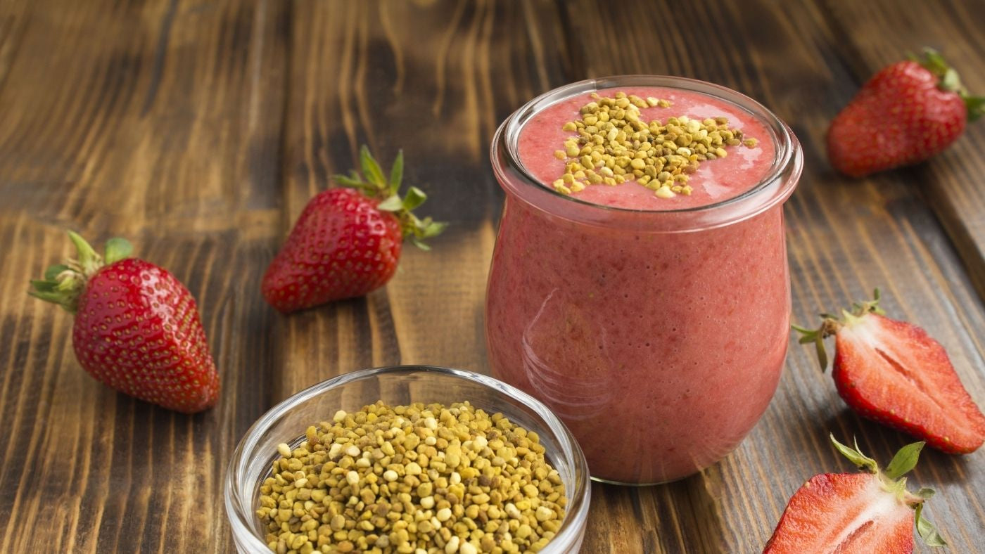 Bee Pollen Granules in a bowl next to Creamy Bee Pollen Strawberry Smoothy in glass surrounded by fresh strawberries