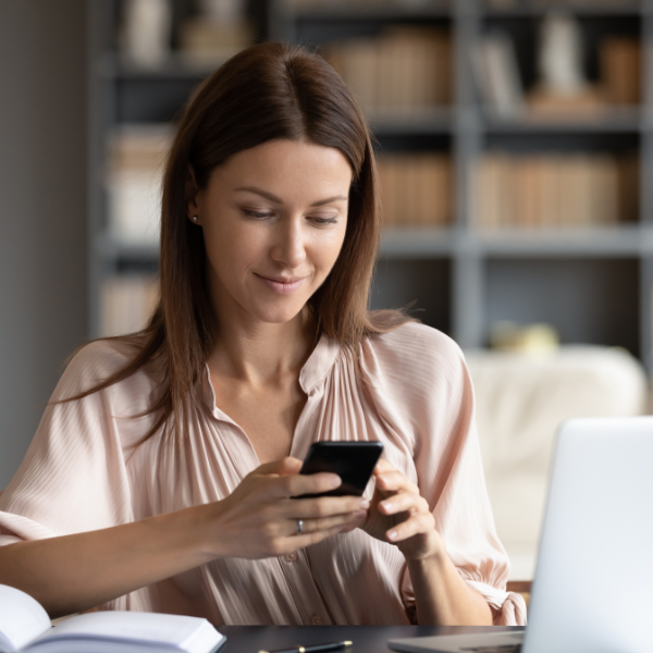 Woman using a smartphone in a home office setting with a laptop and books in the background.
